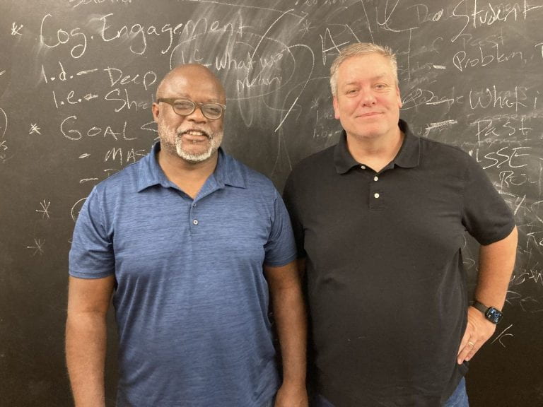two men standing in front of a blackboard with writing on it are smiling